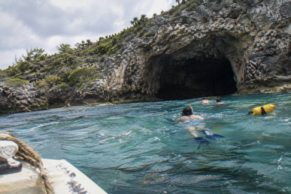 Snorkelers swimming near the entrance of a limestone cave in shallow clear water