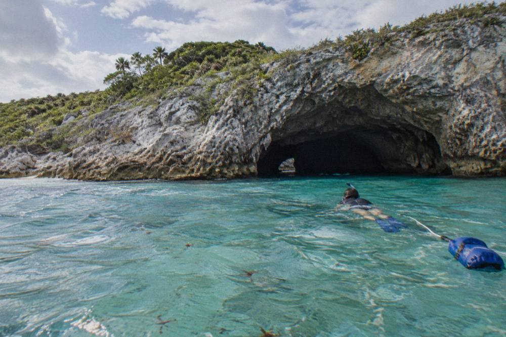 A snorkeler swimming near the entrance of a sea cave above clear shallow water